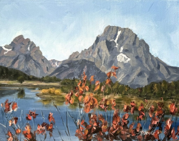 A serene landscape showcases majestic mountains towering over a calm lake with reflections in the water. In the foreground, vibrant red foliage adds a pop of color to the scene suggesting a season change by Mary Benke